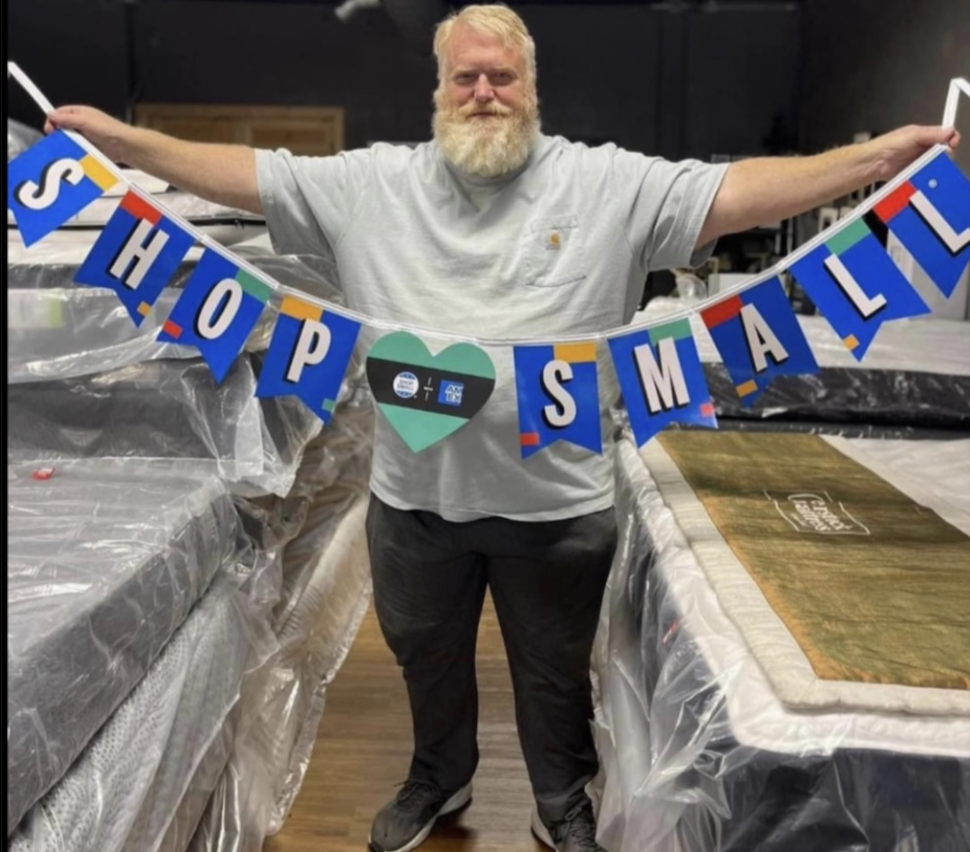 Carey Crowder holding a Shop Small banner inside the Overstock Mattress & Beds showroom